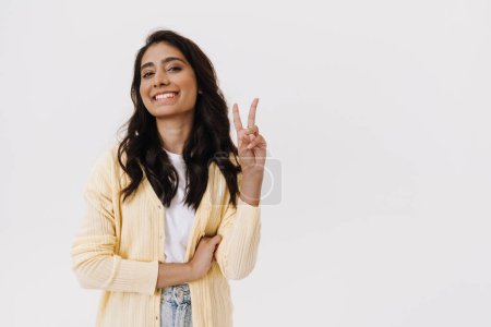 Téléchargez les photos : Jeune femme indienne brune souriant à la caméra et geste signe de paix isolé sur fond blanc - en image libre de droit