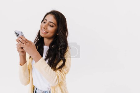 Téléchargez les photos : Jeune femme indienne brune souriante en utilisant un téléphone portable isolé sur fond blanc - en image libre de droit