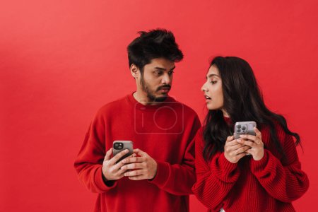 Foto de Joven pareja india asomándose a los celulares del otro mientras posan juntos aislados sobre fondo rojo - Imagen libre de derechos