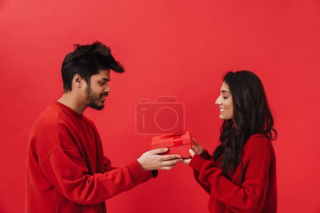 Téléchargez les photos : Jeune homme indien donnant boîte cadeau à sa petite amie isolée sur fond rouge - en image libre de droit