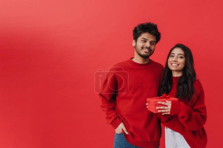 Photo for Young indian couple smiling while posing together with present box isolated over red background - Royalty Free Image