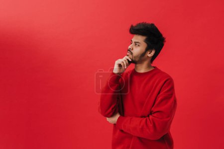 Photo for Young brunette indian man thinking and looking aside while posing at camera isolated over red background - Royalty Free Image