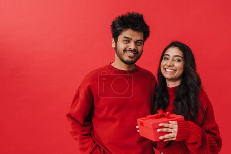 Foto de Joven pareja india sonriendo mientras posan junto con la caja actual aislada sobre fondo rojo - Imagen libre de derechos