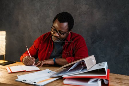 Foto de Hombre barbudo negro en anteojos anotando notas mientras trabajaba en la oficina - Imagen libre de derechos
