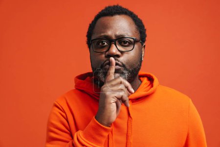 Téléchargez les photos : Homme afro-américain adulte tenant le doigt à la bouche et demandant à garder le silence debout isolé sur fond orange - en image libre de droit