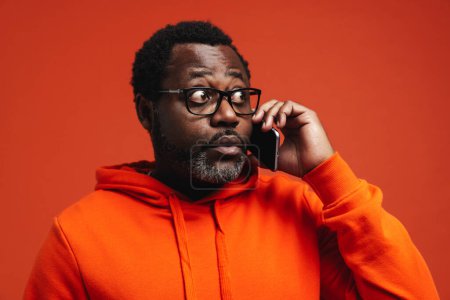Foto de Adulto emocionado hombre americano hablando en el teléfono celular de pie aislado sobre fondo naranja - Imagen libre de derechos