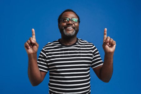 Foto de Hombre afroamericano adulto con anteojos sonriendo y haciendo gestos hacia arriba aislado sobre fondo azul - Imagen libre de derechos