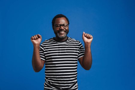 Photo for Adult happy african american man smiling and clenching fists as winner standing isolated over blue background - Royalty Free Image