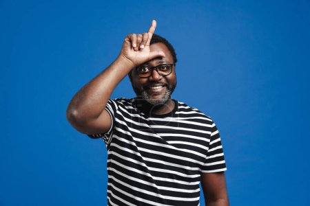 Téléchargez les photos : Adulte heureux homme afro-américain souriant et tenant les doigts à son front debout isolé sur fond bleu - en image libre de droit