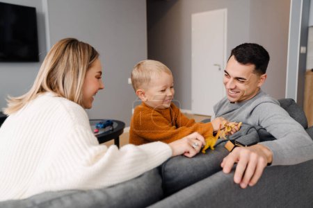 Téléchargez les photos : Heureux parents blancs et petit fils souriant et jouant avec des jouets assis sur le canapé à la maison - en image libre de droit