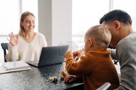 Téléchargez les photos : Jeune femme blanche utilisant un ordinateur portable tandis que l'homme et le petit fils jouent avec des jouets sur la table à la maison - en image libre de droit