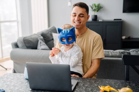 Téléchargez les photos : Jeune homme blanc et petit fils riant et utilisant ordinateur portable tout en étant assis sur la table à la maison - en image libre de droit