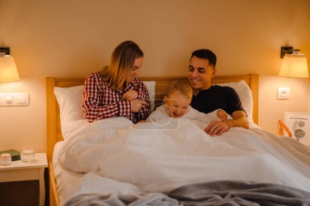 Téléchargez les photos : Jeune homme et femme blancs souriant alors qu'ils étaient couchés au lit avec un petit fils à la maison - en image libre de droit