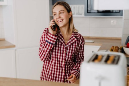 Téléchargez les photos : Jeune femme blanche portant un pyjama debout près de la cuisinière dans la cuisine et parlant sur téléphone portable - en image libre de droit