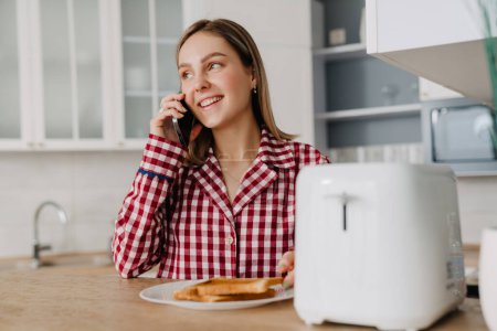Téléchargez les photos : Jeune femme blanche portant un pyjama manger des toasts et parler sur un téléphone portable dans la cuisine - en image libre de droit