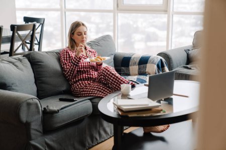 Photo for Young white happy woman wearing pajama having breakfast while sitting on sofa at home - Royalty Free Image