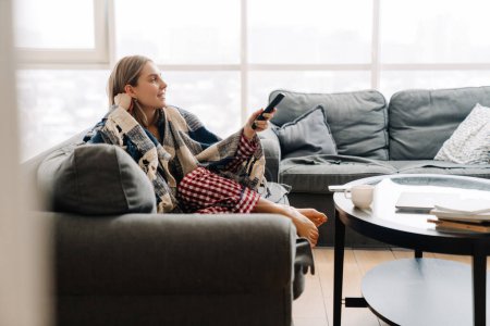 Téléchargez les photos : Jeune femme blanche heureuse enveloppée dans une couverture tenant la télécommande tout en regardant la télévision sur le canapé à la maison - en image libre de droit