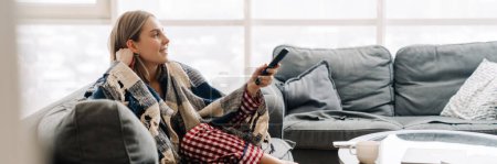 Téléchargez les photos : Jeune femme blanche heureuse enveloppée dans une couverture tenant la télécommande tout en regardant la télévision sur le canapé à la maison - en image libre de droit