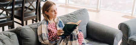 Foto de Joven mujer blanca envuelta en libro de lectura de manta mientras está sentada en el sofá en casa - Imagen libre de derechos