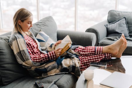 Photo pour Jeune femme blanche heureuse enveloppée dans un livre de lecture de couverture alors qu'elle était assise sur un canapé à la maison - image libre de droit