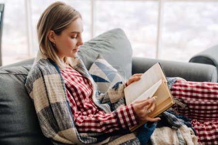 Foto de Joven mujer feliz blanco envuelto en libro de lectura manta mientras está sentado en el sofá en casa - Imagen libre de derechos