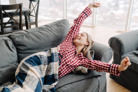 Téléchargez les photos : Jeune femme blanche portant un pyjama souriant alors qu'elle était allongée sur le canapé à la maison le matin - en image libre de droit