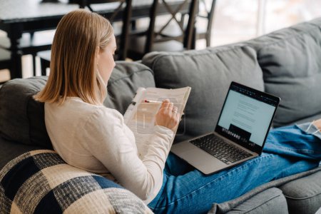 Foto de Mujer rubia blanca joven usando el ordenador portátil mientras estudia con el libro de ejercicios en el sofá en casa - Imagen libre de derechos