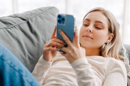 Photo for Young white blond woman using cellphone while lying on sofa at home - Royalty Free Image