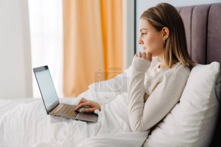 Foto de Mujer rubia blanca joven usando el ordenador portátil mientras está acostado en la cama en casa por la mañana - Imagen libre de derechos