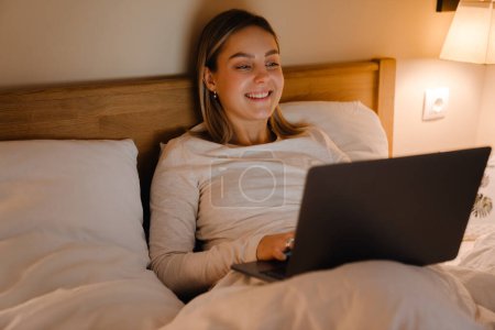 Téléchargez les photos : Jeune femme blanche souriant et utilisant un ordinateur portable tout en étant couché dans le lit à la maison - en image libre de droit