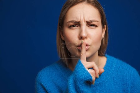 Téléchargez les photos : Jeune femme blanche tendue portant un pull fronçant les sourcils et faisant un geste de silence isolé sur fond bleu - en image libre de droit