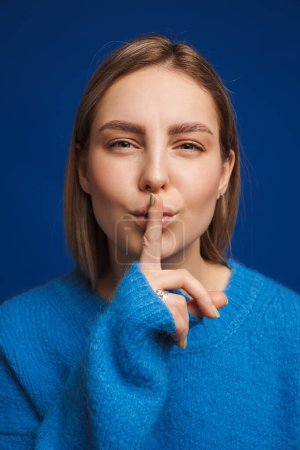 Photo for Portrait of young beautiful girl showing silence gesture looking in camera over blue isolated background - Royalty Free Image
