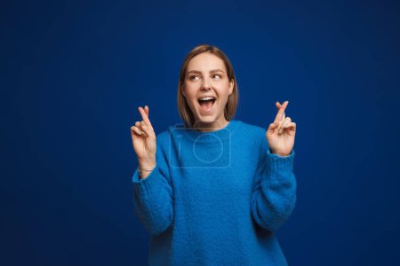Photo for Young hopeful beautiful girl with csossed fingers and opened mouth looking leftward over blue isolated background - Royalty Free Image