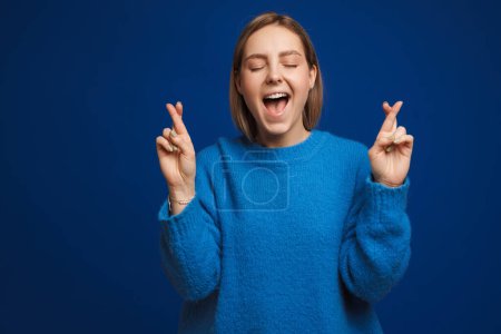 Photo for Young hopeful beautiful girl with csossed fingers, closed eyes and opened mouth over blue isolated background - Royalty Free Image