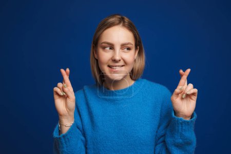 Photo for Young beautiful hopeful girl bite her lips with crossed fingers looking rightward over isolated blue background - Royalty Free Image