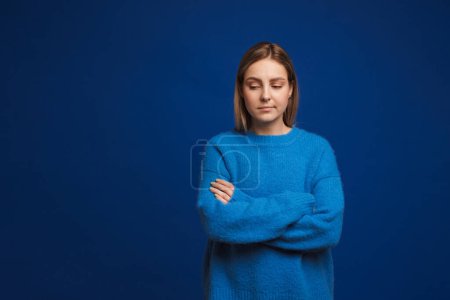 Foto de Joven chica triste con brazos cruzados en suéter azul mirando hacia abajo de pie sobre fondo azul aislado - Imagen libre de derechos