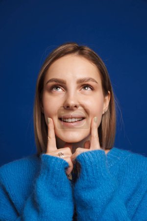 Téléchargez les photos : Jeune femme blanche heureuse portant un pull souriant et touchant son visage debout isolé sur fond bleu - en image libre de droit