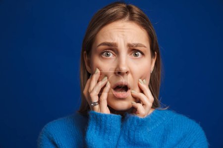 Photo for Young scared girl touching her cheeks with opened mouth over isolated blue background - Royalty Free Image