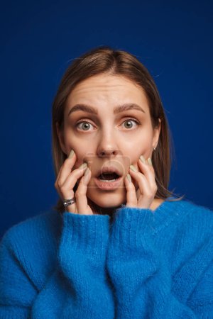 Foto de Joven chica sorprendida tocando sus mejillas con la boca abierta sobre fondo azul aislado - Imagen libre de derechos