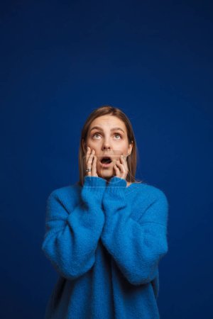 Foto de Joven chica sorprendida tocándose las mejillas con la boca abierta mirando hacia arriba y de pie sobre un fondo azul aislado - Imagen libre de derechos