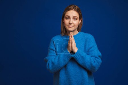 Foto de Joven chica guapa en suéter azul mostrando gesto de oración de pie sobre fondo azul aislado - Imagen libre de derechos
