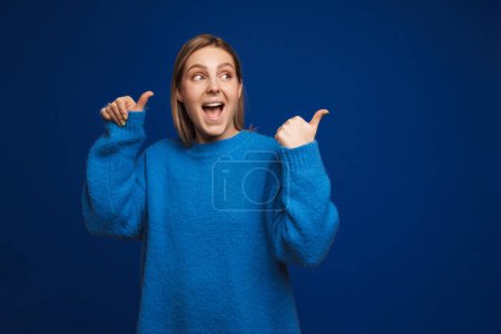 Foto de Joven chica guapa sonriente en suéter azul apuntando y mirando hacia la derecha de pie sobre fondo azul aislado - Imagen libre de derechos