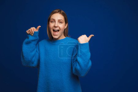 Foto de Joven chica sonriente guapo en suéter azul apuntando hacia la derecha y mirando en la cámara de pie sobre fondo azul aislado - Imagen libre de derechos