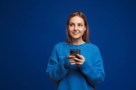 Foto de Joven chica guapa en suéter azul sosteniendo el teléfono y mirando hacia la izquierda de pie sobre fondo azul aislado - Imagen libre de derechos