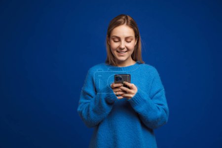 Téléchargez les photos : Jeune belle fille en pull bleu tenant le téléphone et le regardant debout sur fond bleu isolé - en image libre de droit