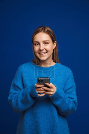 Téléchargez les photos : Jeune belle fille en pull bleu tenant le téléphone et regardant dans la caméra debout sur fond bleu isolé - en image libre de droit