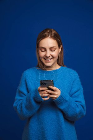 Foto de Joven chica guapa en suéter azul sosteniendo el teléfono y mirándolo de pie sobre fondo azul aislado - Imagen libre de derechos