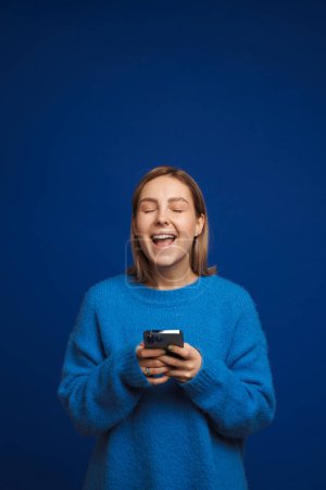 Photo pour Jeune belle fille heureuse tenant le téléphone avec les yeux fermés et la bouche ouverte debout sur fond bleu isolé - image libre de droit