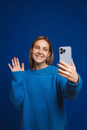 Foto de Joven hermosa chica tomando selfie o haciendo videocall saludando a la cámara sobre aislado fondo azul - Imagen libre de derechos