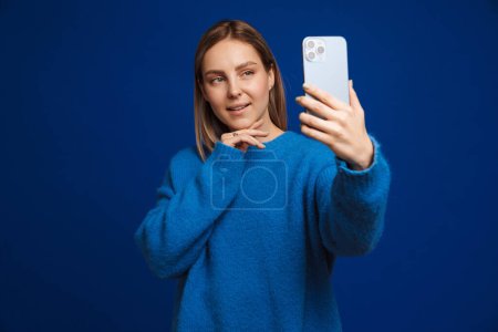 Téléchargez les photos : Jeune belle fille prendre selfie toucher son menton debout sur isolé bleu backround - en image libre de droit